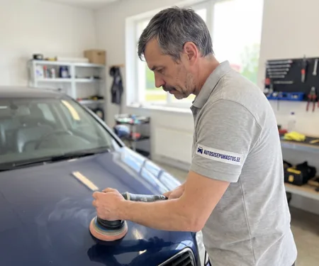 Poleerija töötab oranži padjaga masinaga tumesinise auto kapotil, poleerimismasina juhe hoitakse üle õla, et see üle laki ei lohiseks.